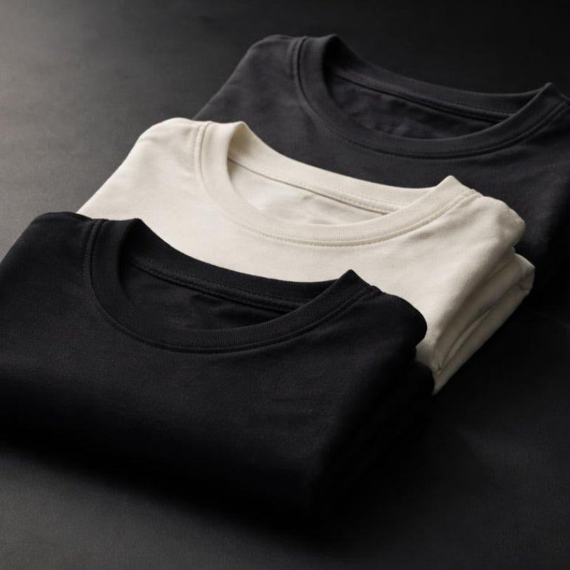 Three folded t-shirts in black, white, and gray on a dark surface